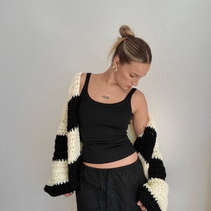 Pode incluir: Um cardigan de croch&ecirc; preto e creme, oversized, com um padr&atilde;o xadrez arrojado. O su&eacute;ter aberto &eacute; usado sobre uma regata preta e cal&ccedil;as pretas. As mangas do cardigan s&atilde;o longas e largas.