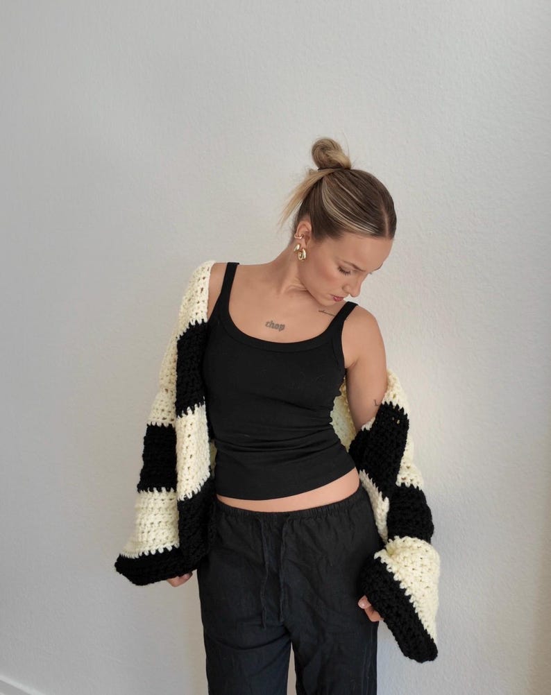 Pode incluir: Um cardigan de croch&ecirc; preto e creme, oversized, com um padr&atilde;o xadrez arrojado. O su&eacute;ter aberto &eacute; usado sobre uma regata preta e cal&ccedil;as pretas. As mangas do cardigan s&atilde;o longas e largas.
