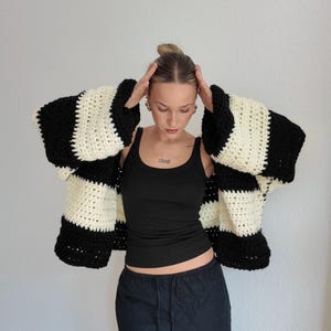 Pode incluir: Um cardigan de croch&ecirc; listrado em preto e creme com mangas grandes. O tric&ocirc; grosso apresenta listras horizontais arrojadas. A modelo veste um top preto e cal&ccedil;as pretas. O cardigan &eacute; uma pe&ccedil;a marcante.