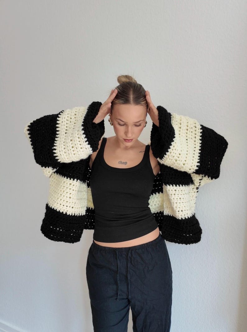 Pode incluir: Um cardigan de croch&ecirc; listrado em preto e creme com mangas grandes. O tric&ocirc; grosso apresenta listras horizontais arrojadas. A modelo veste um top preto e cal&ccedil;as pretas. O cardigan &eacute; uma pe&ccedil;a marcante.