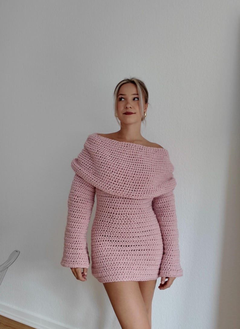 May include: A light pink, off-the-shoulder crochet dress. The dress features long sleeves and a fitted silhouette. The neckline is wide and draped, and the dress appears to be handmade. The overall style is casual and feminine.