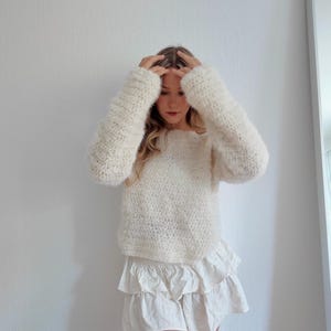 May include: Cream-colored, long-sleeved sweater with a textured knit. The sweater is paired with a white, tiered skirt. The outfit is casual and comfortable, suitable for everyday wear.