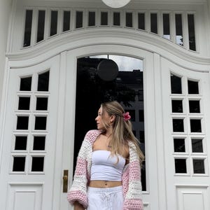 May include: A woman wearing a pink and white striped crocheted cardigan with white pants and a white tank top. She is standing in front of a white door with a black door handle.