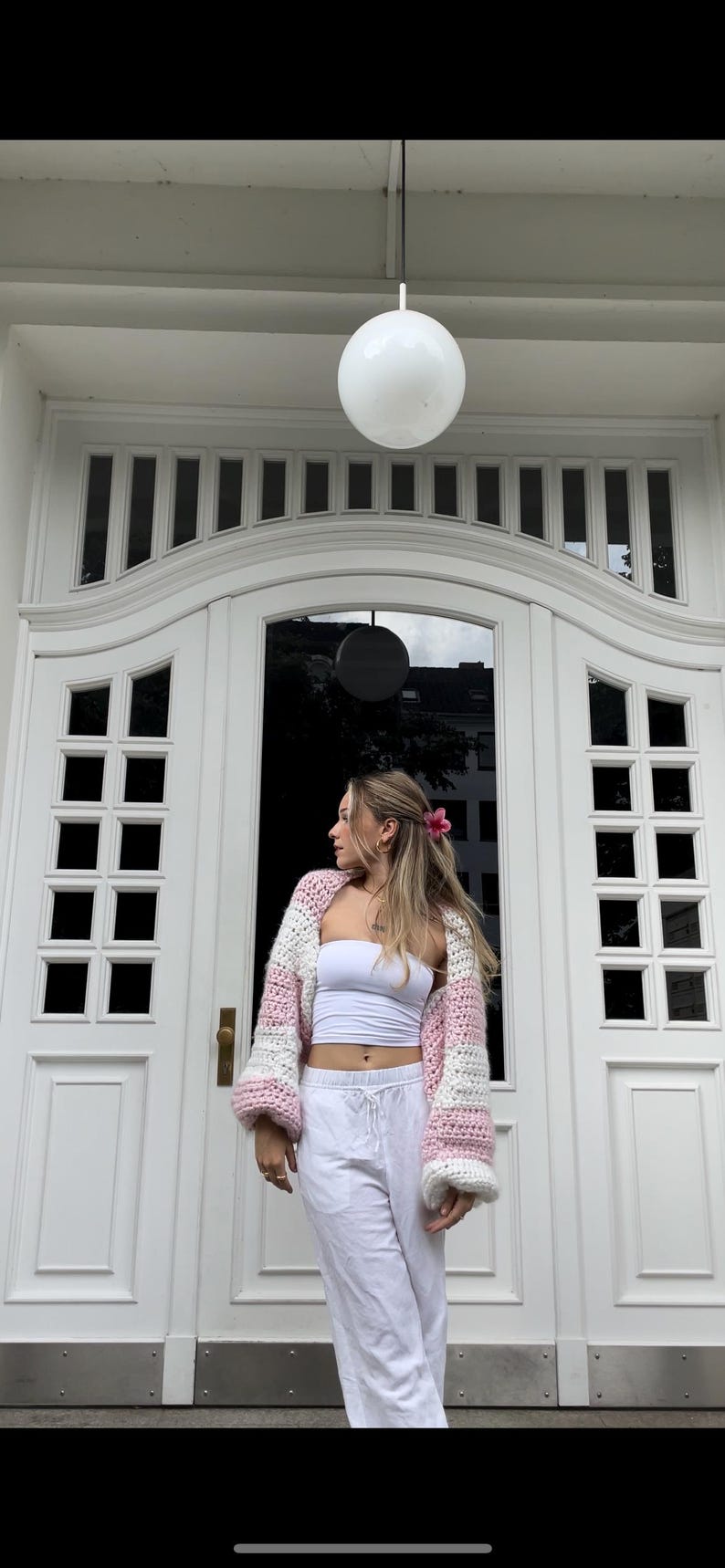 May include: A woman wearing a pink and white striped crocheted cardigan with white pants and a white tank top. She is standing in front of a white door with a black door handle.