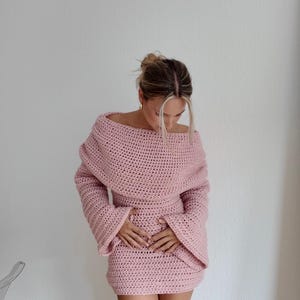 May include: A light pink, off-the-shoulder crochet dress with long sleeves. The dress features a fitted waist and a mini skirt. The model is wearing the dress. The dress is handmade and has a textured appearance.