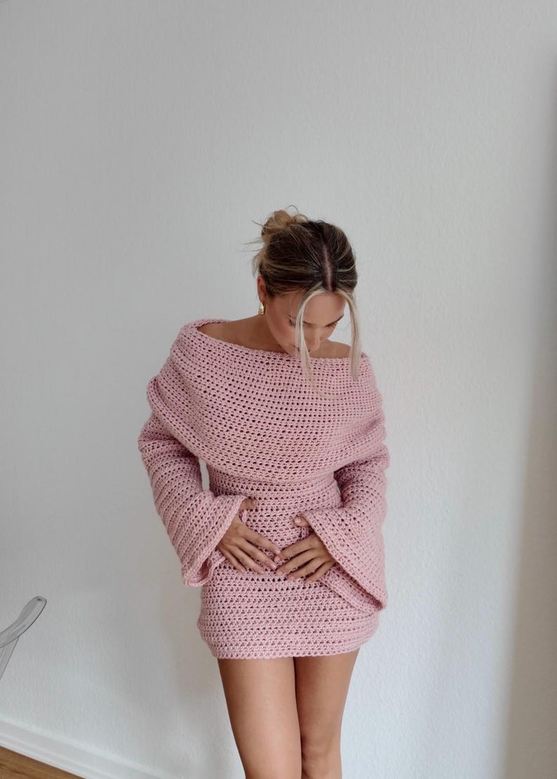 May include: A light pink, off-the-shoulder crochet dress with long sleeves. The dress features a fitted waist and a mini skirt. The model is wearing the dress. The dress is handmade and has a textured appearance.