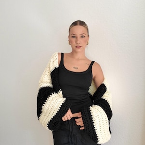 Pode incluir: Uma regata preta e cal&ccedil;as pretas. Um cardigan de croch&ecirc; com mangas largas em listras pretas e creme alternadas &eacute; usado. O cardigan &eacute; uma pe&ccedil;a ousada, com uma combina&ccedil;&atilde;o de cores marcante.