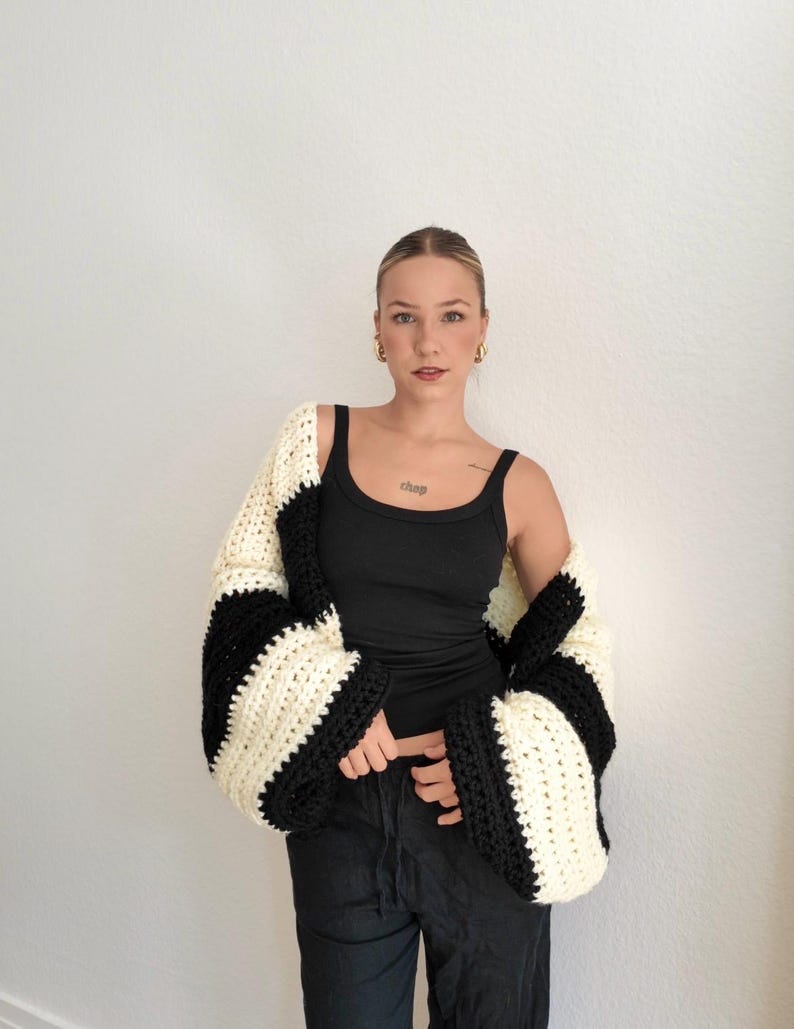 Pode incluir: Uma regata preta e cal&ccedil;as pretas. Um cardigan de croch&ecirc; com mangas largas em listras pretas e creme alternadas &eacute; usado. O cardigan &eacute; uma pe&ccedil;a ousada, com uma combina&ccedil;&atilde;o de cores marcante.