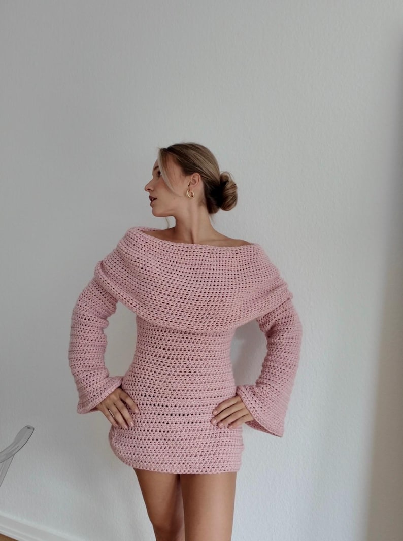 May include: A light pink, off-the-shoulder crochet dress with long sleeves. The dress has a fitted bodice and a flared skirt. The sleeves are bell-shaped. The model is wearing the dress.
