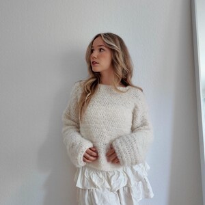 May include: Cream-colored, hand-knitted sweater with wide sleeves and a white ruffled skirt. The sweater has a textured appearance. The outfit is casual and stylish, suitable for everyday wear.