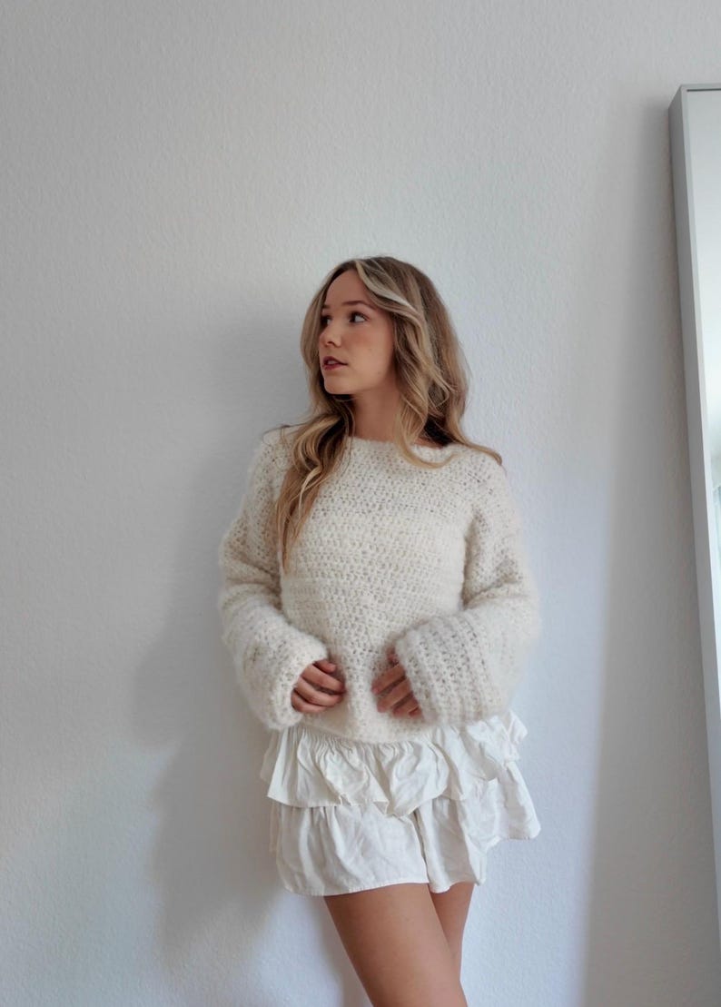May include: Cream-colored, hand-knitted sweater with wide sleeves and a white ruffled skirt. The sweater has a textured appearance. The outfit is casual and stylish, suitable for everyday wear.