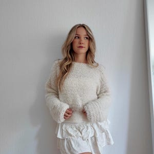 May include: Cream-colored, oversized knit sweater with long, flared sleeves. Paired with a white, tiered ruffle skirt. The outfit is casual and comfortable, suitable for everyday wear. The sweater has a textured appearance.