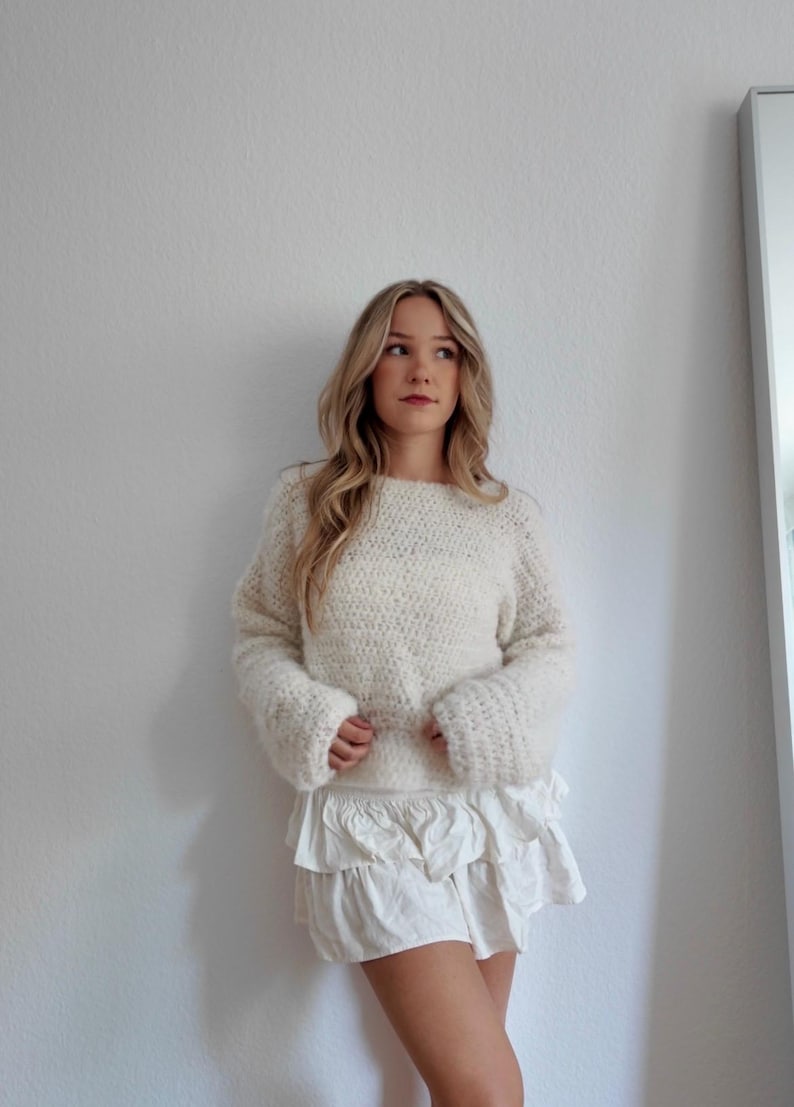 May include: Cream-colored, oversized knit sweater with long, flared sleeves. Paired with a white, tiered ruffle skirt. The outfit is casual and comfortable, suitable for everyday wear. The sweater has a textured appearance.