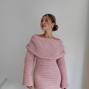 May include: A light pink, off-the-shoulder crochet dress with long sleeves. The dress has a fitted silhouette and a unique, draped neckline. The crochet stitch creates a textured appearance. The dress is a mini length.