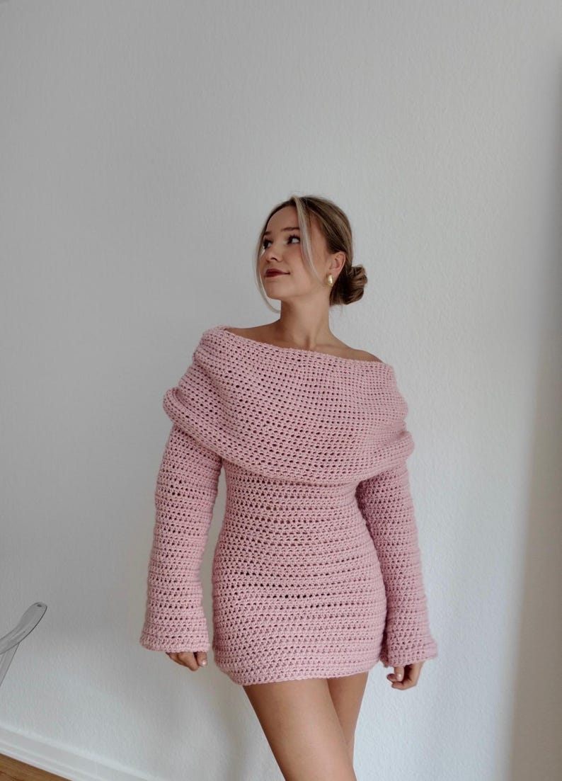 May include: A light pink, off-the-shoulder crochet dress with long sleeves. The dress has a fitted silhouette and a unique, draped neckline. The crochet stitch creates a textured appearance. The dress is a mini length.