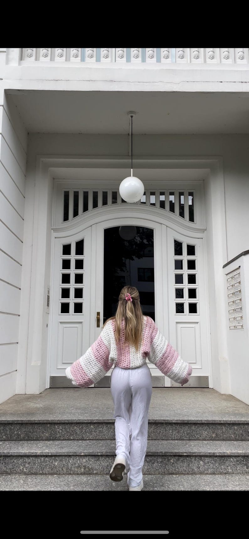 May include: A person wearing a pink and white striped crocheted sweater and white pants, standing on a set of stairs in front of a white door.