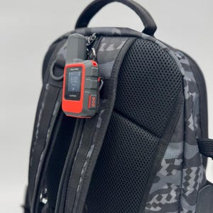 May include: A gray and red Garmin inReach Mini 2 GPS device attached to a black camouflage backpack. The device displays the time as 6:02 and has an SOS button. The backpack has a black mesh back and a top handle.