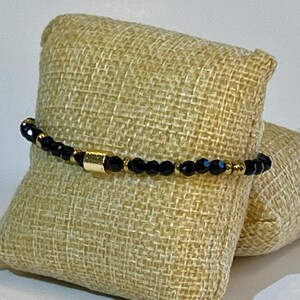 Puede incluir: Un brazalete de cuentas negras con un acento dorado. El brazalete se muestra sobre una superficie de tela beige.