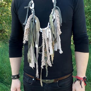 Puede incluir: Un collar hecho a mano con tiras de tela en tonos verdes, beige y grises. El collar tiene un diseño bohemio único con una mezcla de texturas y materiales. El collar se lleva sobre una camiseta negra de manga larga.