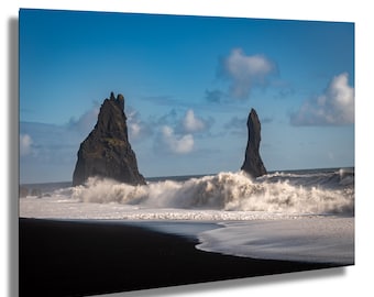 May include: A landscape photograph showing a black sand beach with two large rock formations in the sea. White waves crash against the shore. The sky is blue with scattered clouds, creating a dramatic coastal scene.