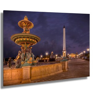 May include: A nighttime view of the Place de la Concorde in Paris, France. The image features the ornate Fontaine des Mers, a tall obelisk, and streetlights against a dark blue sky. The fountain has detailed sculptures and a gold-colored top.