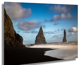 May include: A landscape photograph showing a black sand beach with sea stacks and waves. The sky is blue with clouds. The sea stacks are dark, and the waves are white as they crash on the shore.