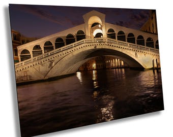 May include: A view of a stone bridge over a canal in Venice, Italy at night. The bridge is lit up with warm yellow lights, and the water reflects the lights. The bridge is arched and has a series of small arches along the sides.