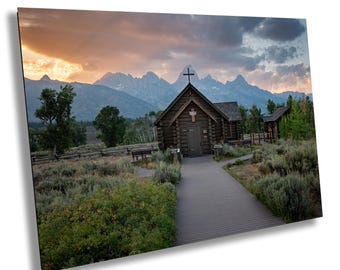May include: A wooden chapel with a cross on the roof sits in front of a mountain range. The chapel is surrounded by green grass and trees. The sky is a soft pink and blue.