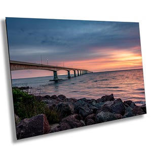 Op de afbeelding: Een lange brug strekt zich uit over het water richting een kleurrijke zonsondergang. De brug heeft meerdere bogen en is gemaakt van beton. Het water is kalm en reflecteert de kleuren van de lucht. Op de voorgrond liggen rotsen.