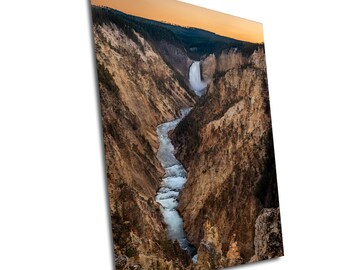 May include: A scenic view of a waterfall cascading down a rocky canyon in Yellowstone National Park. The water flows through a narrow riverbed, surrounded by brown cliffs and lush green trees. The sky is a vibrant orange and yellow, creating a beautiful sunset backdrop.