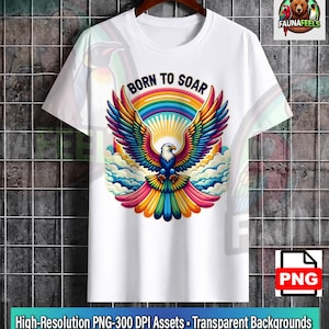 Puede incluir: Una camiseta blanca de manga corta con un gráfico de un águila calva con las alas multicolores extendidas. Sobre el águila, el texto BORN TO SOAR está impreso en semicírculo. El diseño incluye un arcoíris, un sol y nubes.