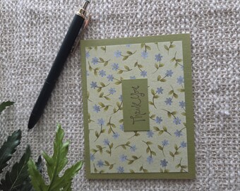 Puede incluir: Una tarjeta de felicitación rectangular con fondo verde claro y un patrón repetitivo de pequeñas flores azules y hojas verdes. Una etiqueta rectangular verde oliva centrada muestra el texto manuscrito Thank You.