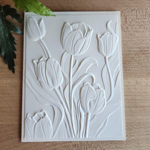 Puede incluir: Escultura en relieve rectangular blanquecina que representa tulipanes. La obra de arte muestra varios tulipanes en diferentes etapas de floración, con pétalos y tallos texturizados. La pieza está sobre un fondo liso, resaltando el diseño floral.