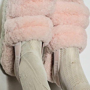 Puede incluir: Zapatillas rosas y esponjosas con suela gris claro. Las zapatillas tienen una textura suave y afelpada y se usan con calcetines beige claro. Una opción de calzado cómoda y acogedora.