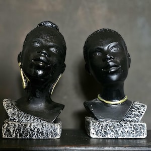 May include: Two black busts of women with detailed facial features. Each bust is mounted on a textured, rectangular base. One wears gold earrings, the other a gold necklace. The busts are displayed on a dark surface against a neutral background.