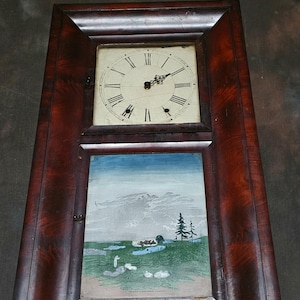 Könnte beinhalten: Antike Wanduhr aus Holz mit rechteckigem Rahmen. Das Zifferblatt hat römische Ziffern und schwarze Zeiger. Unter dem Zifferblatt befindet sich ein Landschaftsbild mit blauem Himmel, grünem Gras und Wasser.