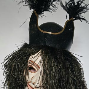 Könnte beinhalten: Eine Theatermaske mit einem schwarzen Helm, der mit schwarzen Federn verziert ist. Die Maske hat ein bemaltes Gesicht mit einem breiten Lächeln und langen, dunklen Haaren. Die Maske wird wahrscheinlich für Kostüm- oder Aufführungszwecke verwendet.
