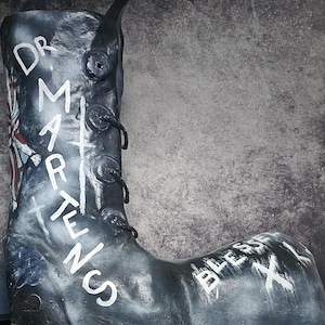 May include: A large, black, sculptural boot with a textured finish. The text "DR MARTENS" is painted in white on the side, with "BLESSED XL" near the toe. A partial Union Jack flag design is visible on the left side, and black laces are present.