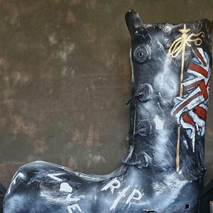 Op de afbeelding: Een grote, zwarte laarssculptuur met witte letters "LOVE" en "RIP". De laars heeft een Union Jack vlagontwerp en gouden accenten. Een uniek decoratief item.