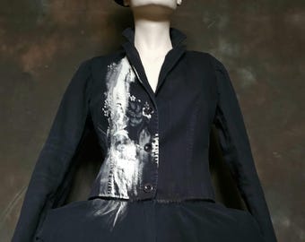 Puede incluir: Un conjunto negro con sombrero de copa, chaqueta y falda, exhibido en un maniquí. La chaqueta tiene un diseño pintado en blanco y negro. La falda tiene un diseño con volantes en capas.
