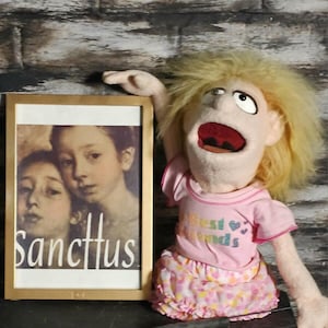 May include: A puppet with blonde hair and a pink shirt and skirt, with the text "Best Friends". Beside the puppet is a framed picture with the word "Sanctus" on it.