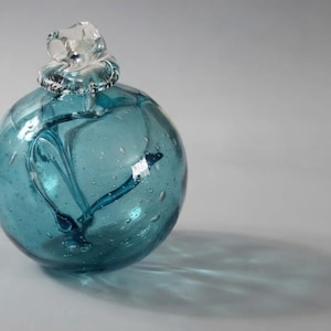 May include: A spherical glass paperweight in a deep teal blue color featuring internal air bubbles and swirling patterns. The top of the orb is finished with a clear, sculpted glass topper that resembles a small, stylized flower or knot.
