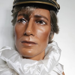 Pode incluir: Uma cabeça de manequim usando um chapéu preto com acabamento dourado e um emblema dourado. O manequim tem uma gola branca com babados. O chapéu tem um pompom marrom no topo.