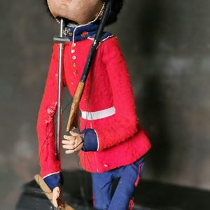 May include: A vintage puppet of a soldier wearing a red coat, black hat, and blue pants. The puppet is holding a wooden rifle and is posed in a standing position.