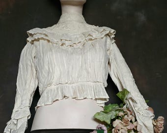 May include: A cream-coloured, ruffled blouse with long sleeves and a high collar is displayed on a mannequin. A black top hat sits atop the mannequin's head. Dried flowers rest on a black, decorative stand, creating a vintage aesthetic.