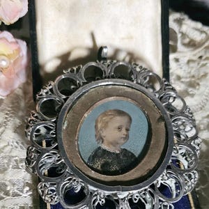 May include: A round silver pendant featuring an intricate filigree border and a central miniature portrait of a child with blonde hair wearing a dark garment. The pendant is displayed inside a black hinged box with a cream-colored interior lining.