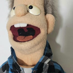 May include: A handmade puppet with a light skin tone, gray hair, and large, expressive eyes. The puppet is wearing a blue and black plaid shirt over a white shirt. The mouth is open, revealing teeth and a red interior.