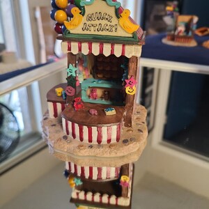May include: A whimsical, multi-tiered decorative figurine resembling a shop. The top features the text "QUACK ATTACK!" and duck figures. The structure is adorned with colorful balloons, smiley faces, and various miniature items.