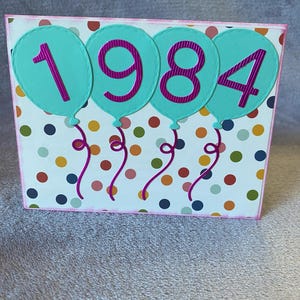 May include: A birthday card with a white background and colorful polka dots. The card features four turquoise balloons with pink numbers spelling out "1984".
