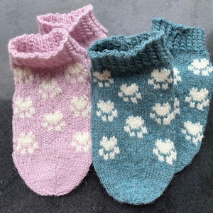 Puede incluir: Dos pares de calcetines de punto con un patrón de huellas de patas. Un par es rosa con huellas de patas blancas y el otro par es azul con huellas de patas blancas.